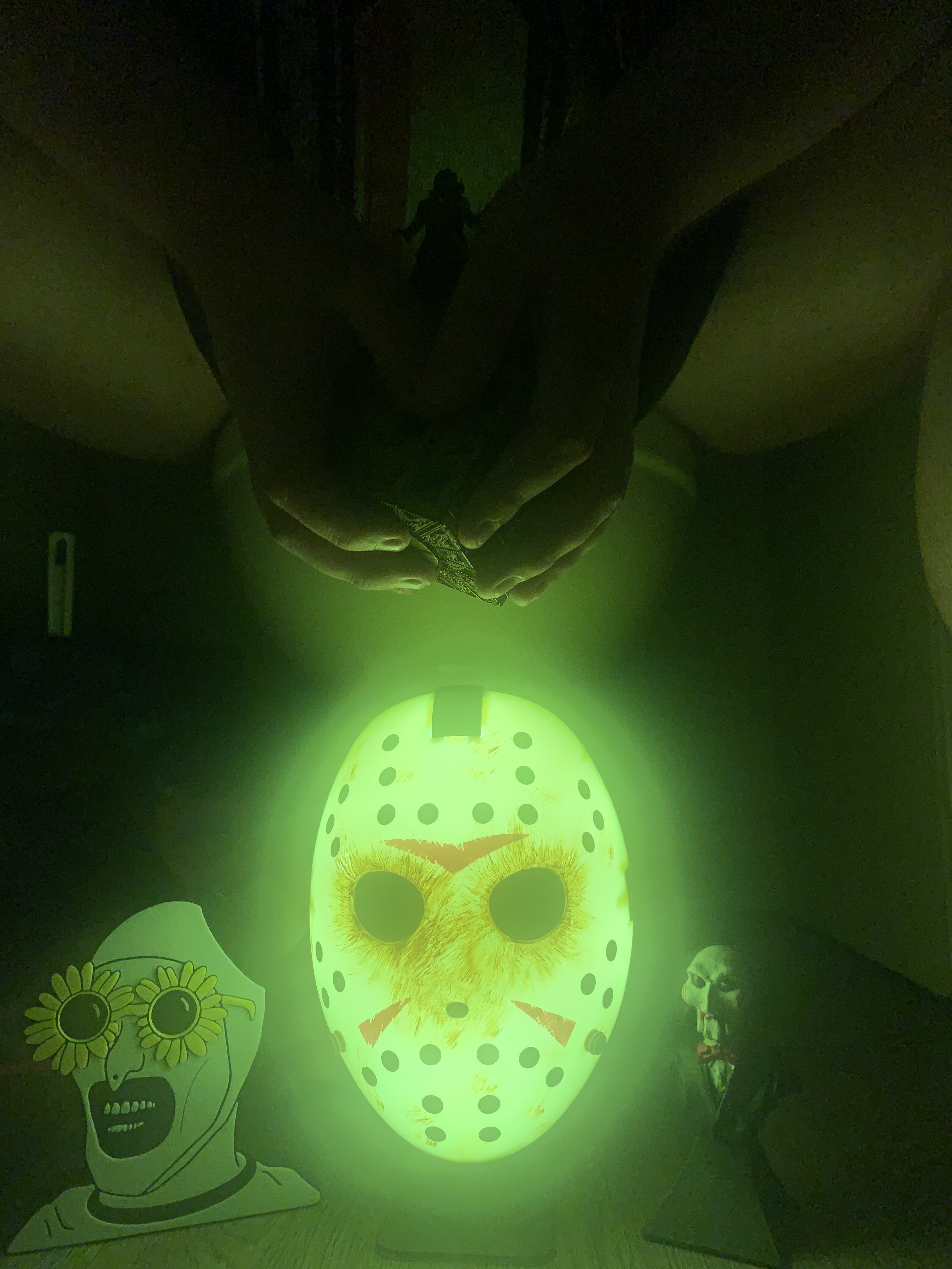 A recreated photo of Hot Wheels & High Heels' banner. A person sits open-legged on a toilet in a dark room, hands brought together at center. Below the hands, a Jason Voorhees mask from Friday the 13th glows green. To the right, a figurine from the Saw franchise. To the left, a character with flower-shaped glasses, grinning with all teeth, wearing what looks like a swimming cap. In the hands rests a Hellraiser puzzle cube, with a small character silhouette balanced on top. The scene is lit by green glow against the dark.