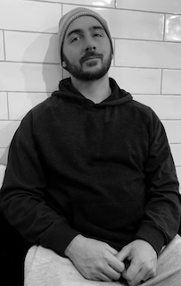 Black and White photo of Jon in a hoodie and beanie against a tiled wall.
